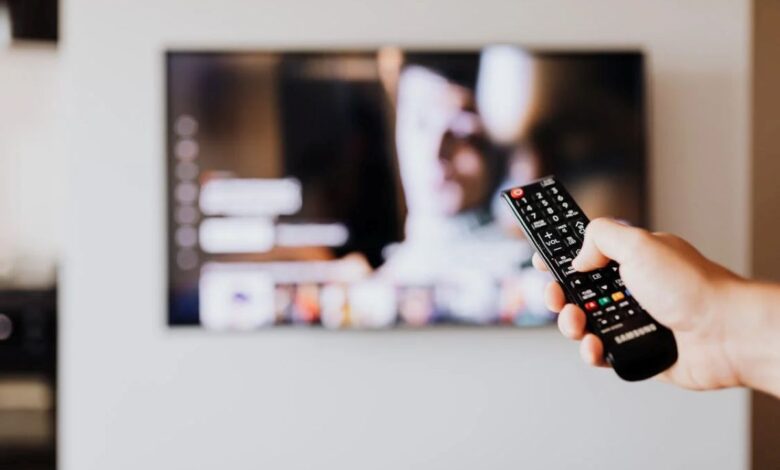 Mão segurando o controle remoto de TV enquanto assiste a uma série ou novela turca, com imagem na tela de um serviço de streaming.