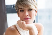 Mulher com corte pixie moderno, sorrindo e inclinando o rosto sobre a mão, mostrando estilo e confiança.