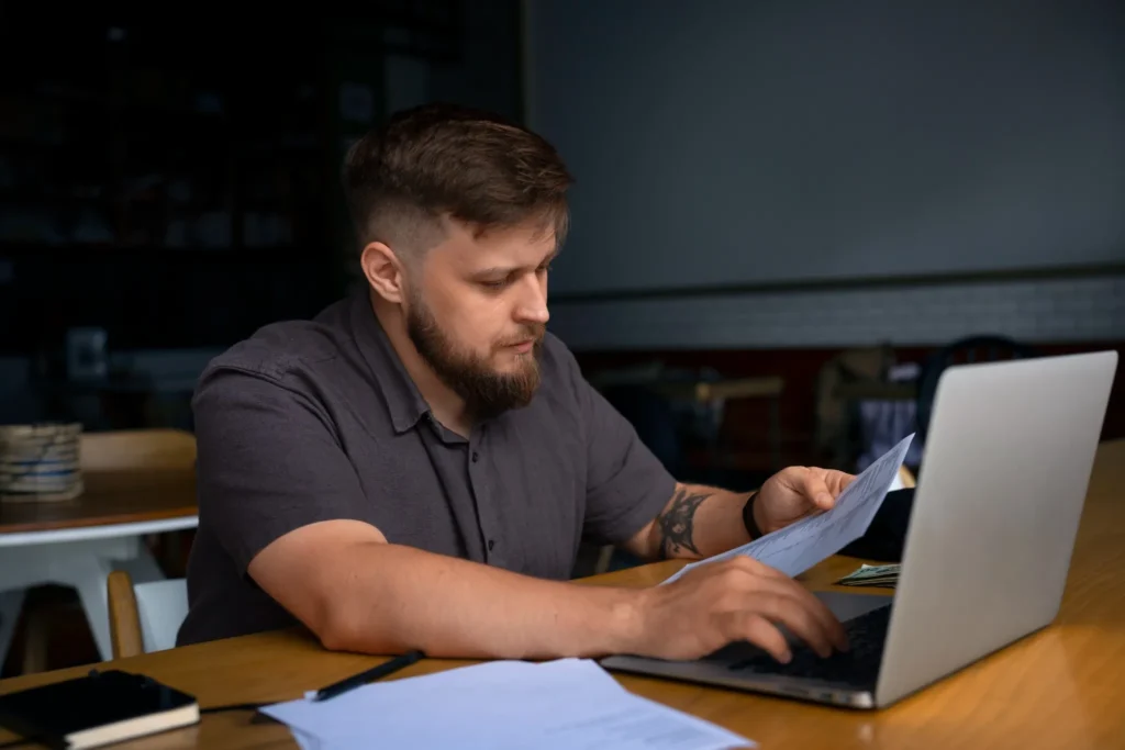 Homem concentrado revisando documentos e usando notebook em ambiente de trabalho
