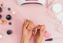 Mãos femininas fazendo manicure com esmalte nude, com ferramentas de manicure ao redor sobre superfície rosa.