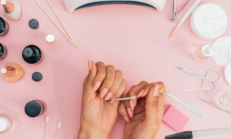 Mãos femininas fazendo manicure com esmalte nude, com ferramentas de manicure ao redor sobre superfície rosa.