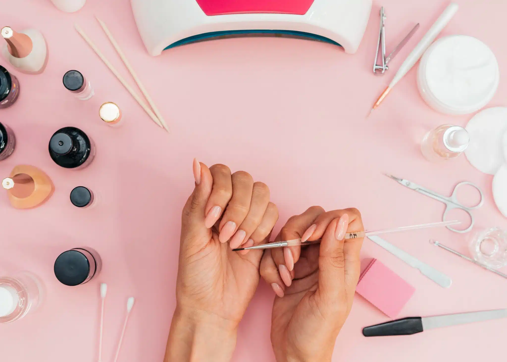 Mãos femininas fazendo manicure com esmalte nude, com ferramentas de manicure ao redor sobre superfície rosa.