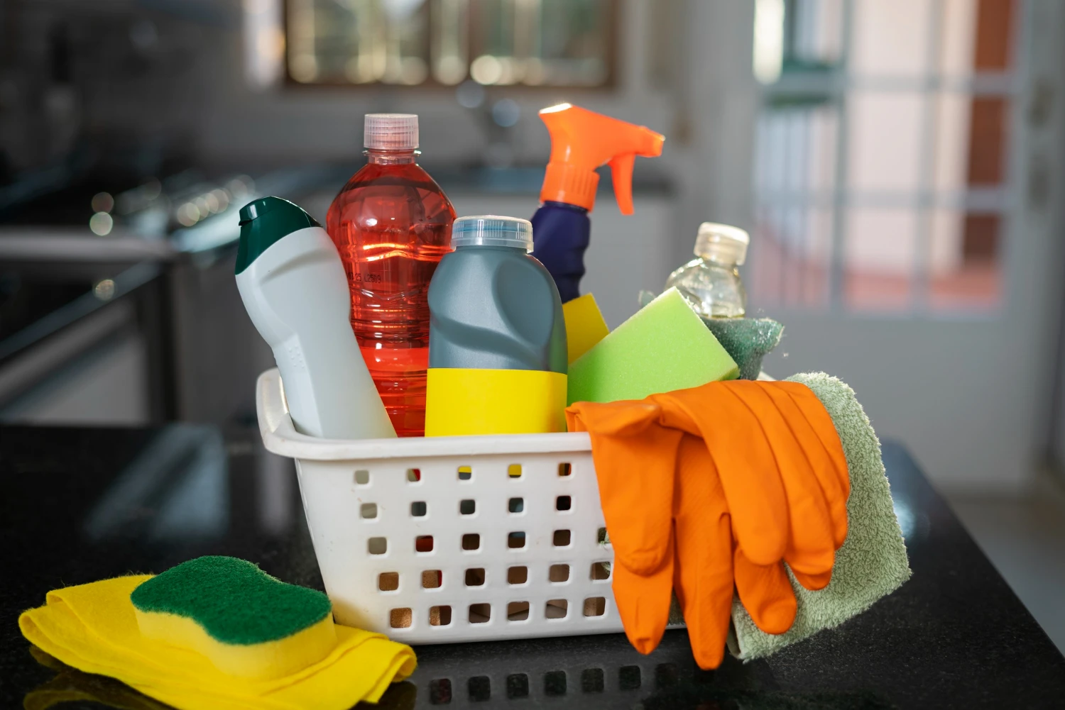 Produtos de limpeza domésticos variados em uma cesta, incluindo esponjas, luvas e frascos.