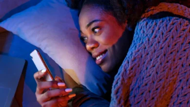 Mulher sorrindo no escuro enquanto segura o celular, envolta em um cobertor, simbolizando uma noite aconchegante e abençoada.