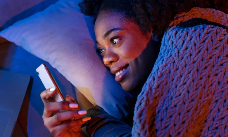 Mulher sorrindo no escuro enquanto segura o celular, envolta em um cobertor, simbolizando uma noite aconchegante e abençoada.