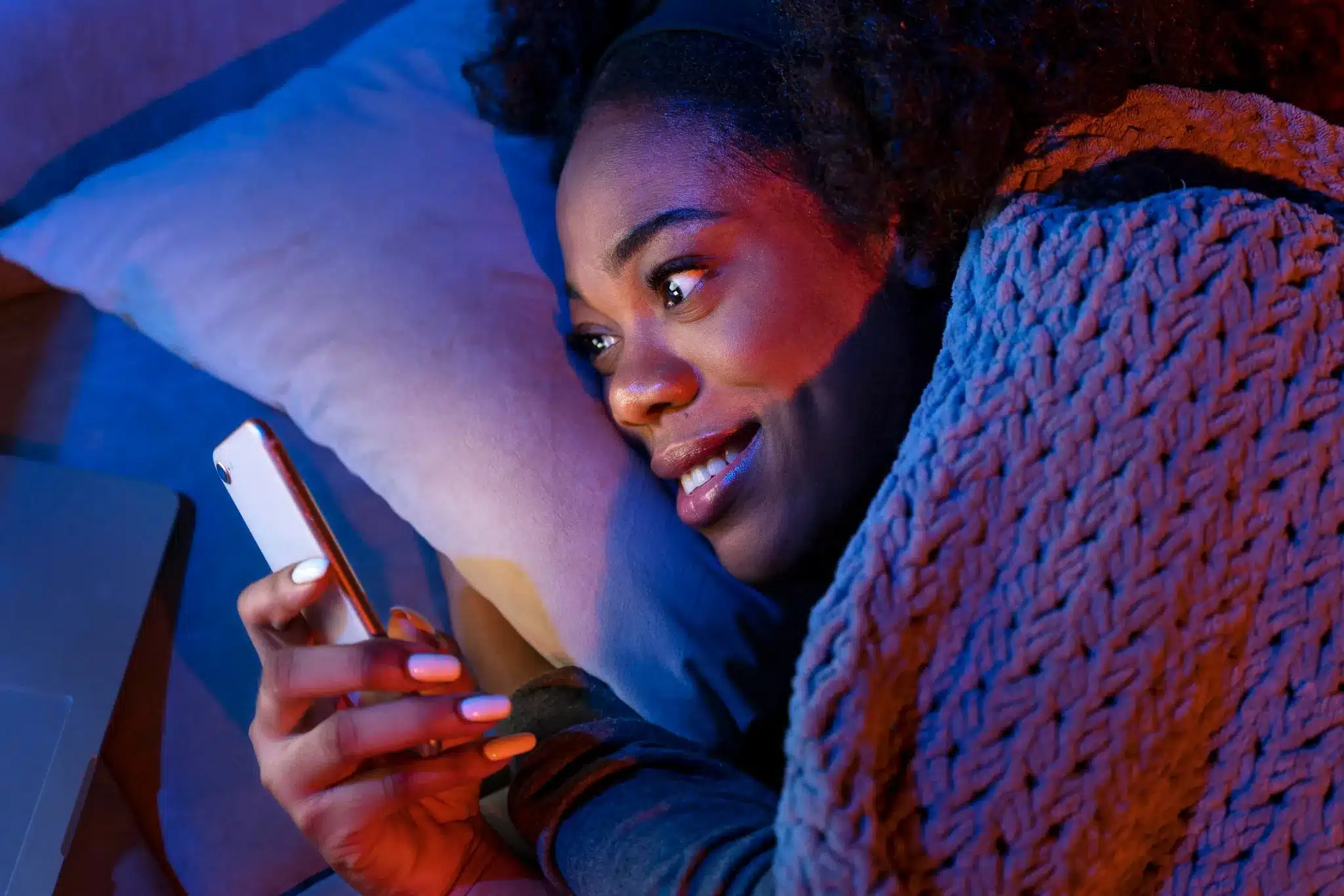 Mulher sorrindo no escuro enquanto segura o celular, envolta em um cobertor, simbolizando uma noite aconchegante e abençoada.