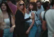Cena da novela turca ‘De Quem Estamos Fugindo’, com duas mulheres se destacando em uma multidão, uma delas com óculos escuros e a outra com cabelo curto e franja, ambas com expressões tensas e intensas