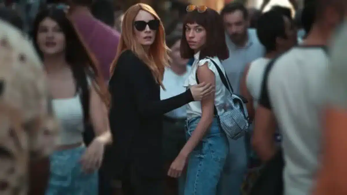 Cena da novela turca ‘De Quem Estamos Fugindo’, com duas mulheres se destacando em uma multidão, uma delas com óculos escuros e a outra com cabelo curto e franja, ambas com expressões tensas e intensas