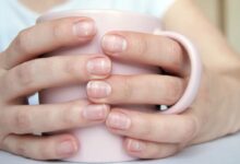 Mãos femininas segurando uma caneca rosa, com unhas naturais mostrando manchas brancas.