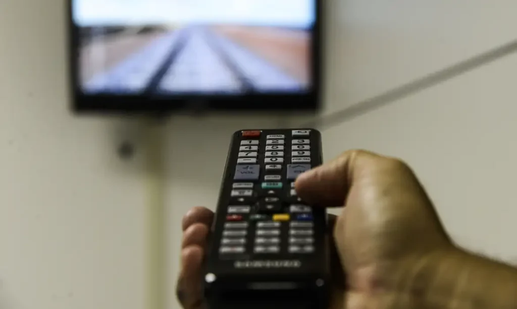 Pessoa segurando controle remoto com TV ligada ao fundo, representando risco de perder o sinal sem a nova parabólica digital