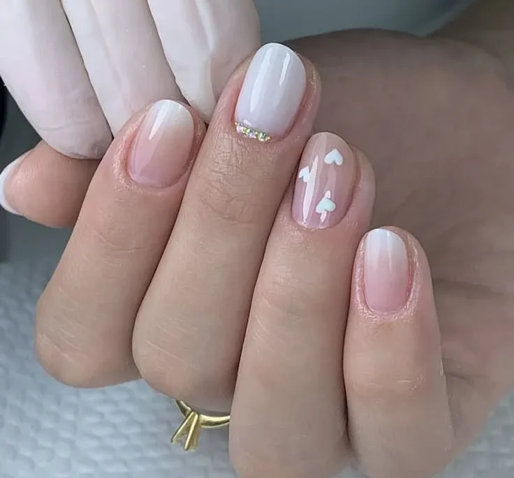 Unhas curtas com formato amendoadas, esmalte nude com detalhes delicados em branco e pequenos strass.