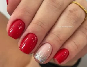 Unhas curtas com esmalte vermelho brilhante e uma unha decorada com glitter prateado.