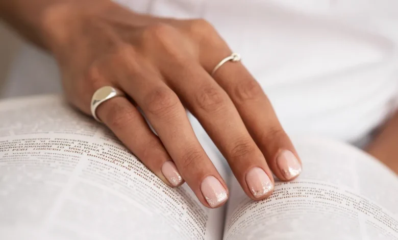 MĂŁo feminina com francesinha delicada em tom nude e glitter, segurando um livro aberto.