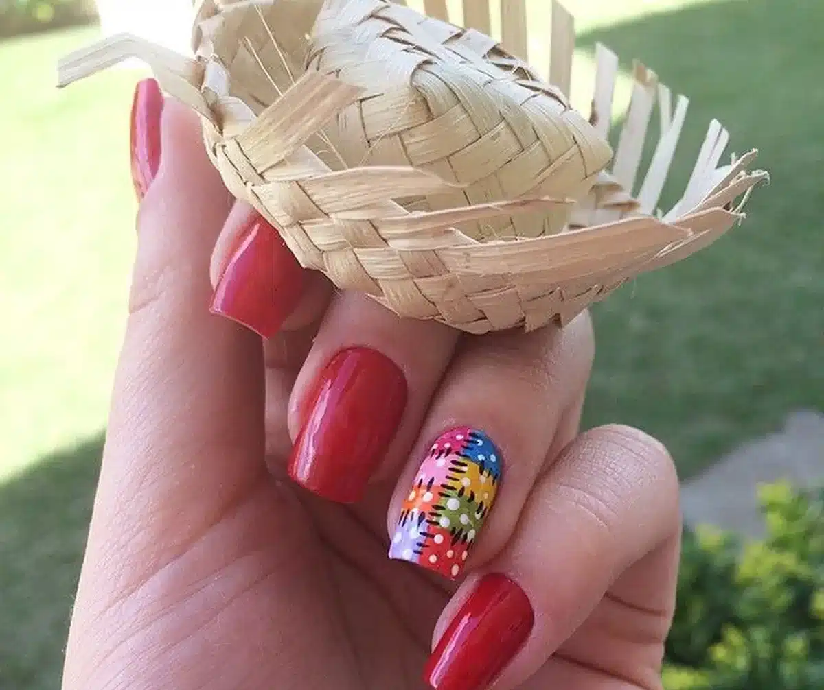 Unhas vermelhas com unha decorada em colorida segurando um mini chapéu de palha