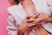 Mulher elegante exibindo unhas postiças longas em tom rosa, com blazer claro e acessórios dourados.