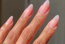 Unhas com esmalte em degradê rosa suave, formato amendoado, adornadas com anéis dourados minimalistas.