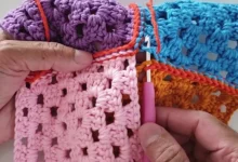 Mãos segurando peças coloridas de crochê unidas com ponto delicado usando agulha