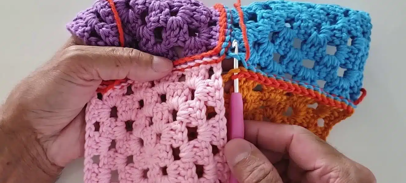 Mãos segurando peças coloridas de crochê unidas com ponto delicado usando agulha