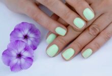 Unhas curtas pintadas de verde leitoso ao lado de flores roxas
