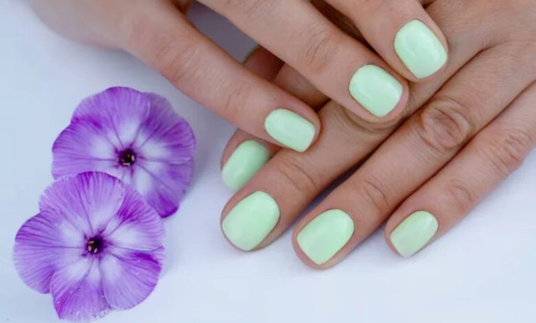 Unhas curtas pintadas de verde leitoso ao lado de flores roxas