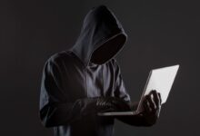 Pessoa encapuzada digitando em laptop em ambiente escuro, representando golpe digital