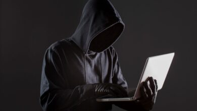 Pessoa encapuzada digitando em laptop em ambiente escuro, representando golpe digital