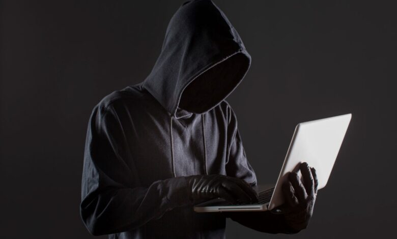 Pessoa encapuzada digitando em laptop em ambiente escuro, representando golpe digital