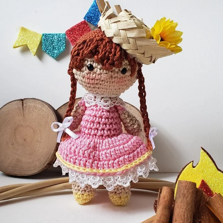 Boneca de amigurumi caipira com vestido rosa e chapéu de palha com flor, perfeita para decoração de festas temáticas.
