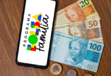 Aplicativo do Bolsa Família ao lado de cédulas e moedas brasileiras sobre mesa de madeira