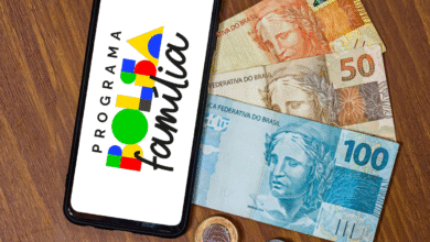 Aplicativo do Bolsa Família ao lado de cédulas e moedas brasileiras sobre mesa de madeira