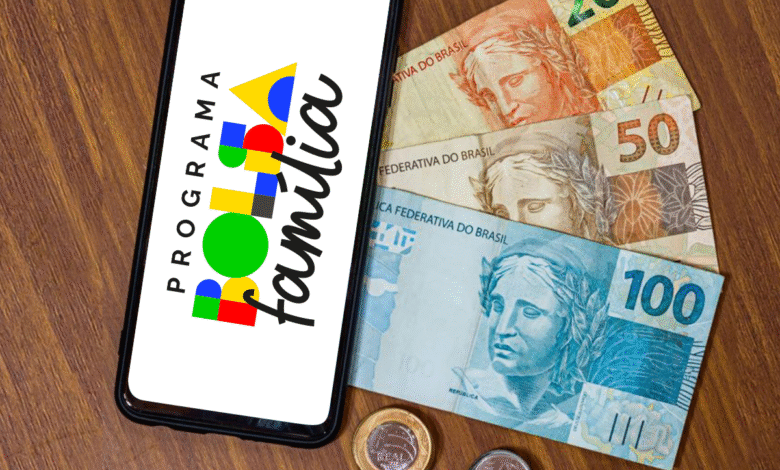 Aplicativo do Bolsa Família ao lado de cédulas e moedas brasileiras sobre mesa de madeira