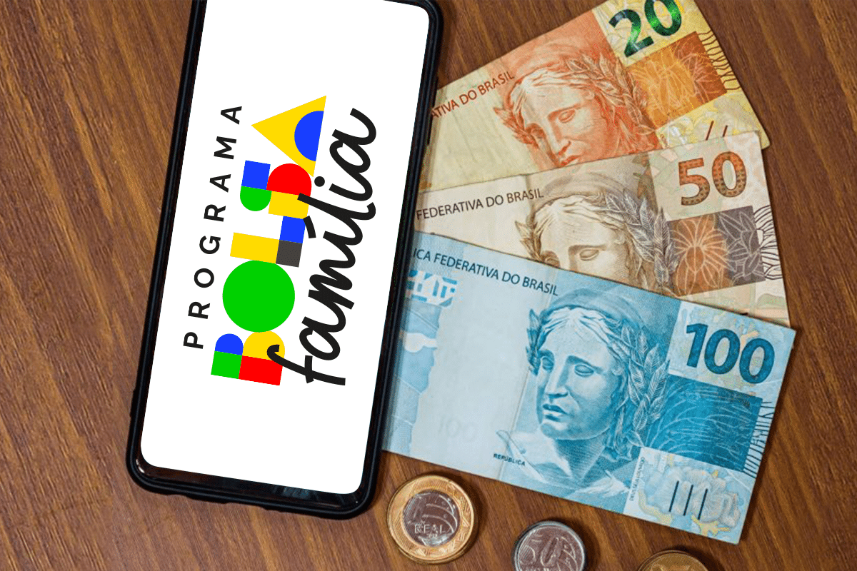 Aplicativo do Bolsa Família ao lado de cédulas e moedas brasileiras sobre mesa de madeira