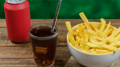 Copo de refrigerante com canudo ao lado de uma tigela cheia de batatas fritas e uma lata vermelha sobre mesa de madeira