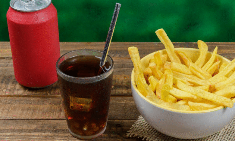 Copo de refrigerante com canudo ao lado de uma tigela cheia de batatas fritas e uma lata vermelha sobre mesa de madeira