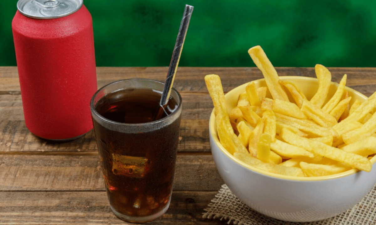 Copo de refrigerante com canudo ao lado de uma tigela cheia de batatas fritas e uma lata vermelha sobre mesa de madeira