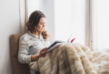 Mulher lendo um livro enquanto está sentada perto da janela com uma manta aconchegante