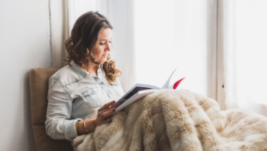 Mulher lendo um livro enquanto está sentada perto da janela com uma manta aconchegante