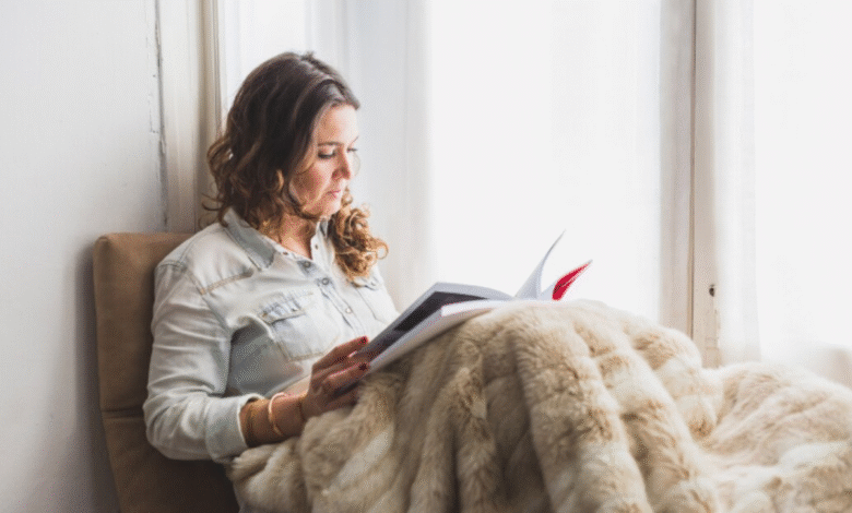 Mulher lendo um livro enquanto está sentada perto da janela com uma manta aconchegante