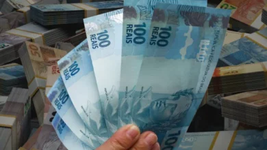 Notas de R$ 100 reais com pilhas de cédulas de diferentes valores ao fundo, ilustrando a grande quantia de dinheiro esquecida nos bancos.