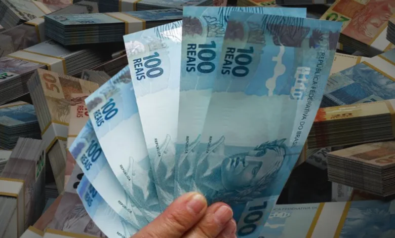 Notas de R$ 100 reais com pilhas de cédulas de diferentes valores ao fundo, ilustrando a grande quantia de dinheiro esquecida nos bancos.