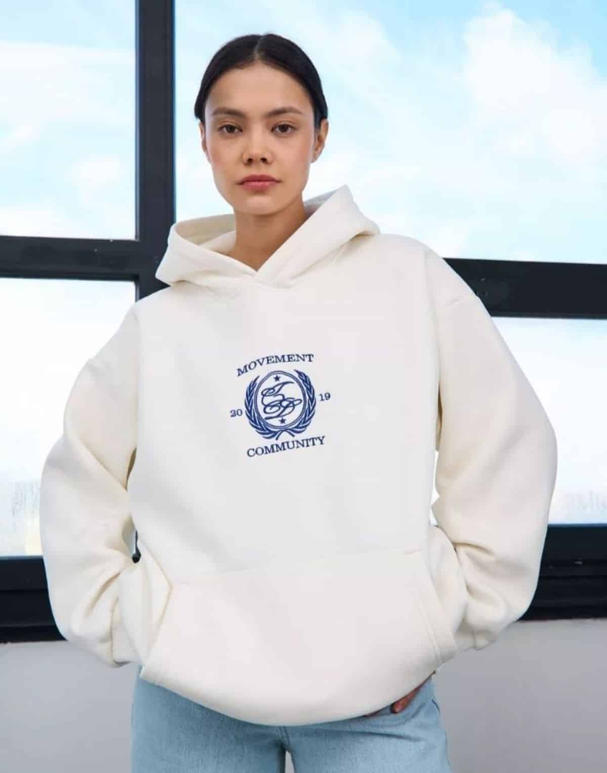 Mulher usando moletom oversized branco com capô e logo, posando em ambiente iluminado