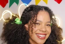 Mulher jovem com cabelos cacheados e laços verdes, sorrindo em ambiente colorido com bandeirinhas juninas no fundo.