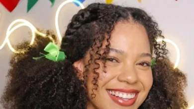 Mulher jovem com cabelos cacheados e laços verdes, sorrindo em ambiente colorido com bandeirinhas juninas no fundo.