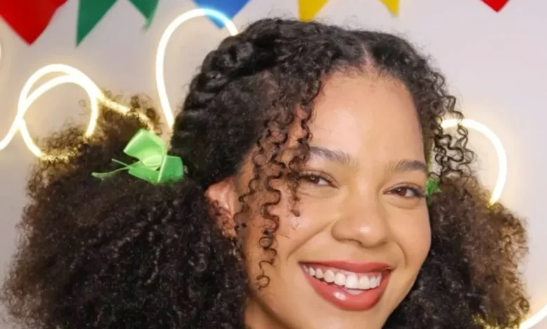 Mulher jovem com cabelos cacheados e laços verdes, sorrindo em ambiente colorido com bandeirinhas juninas no fundo.