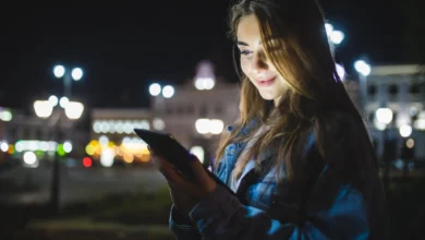 Mulher feliz usando celular sob as luzes da cidade à noite, em clima de boa noite.