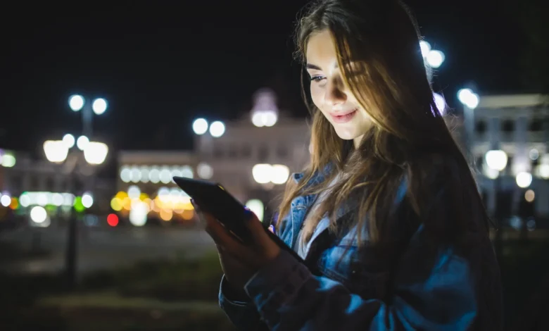 Mulher feliz usando celular sob as luzes da cidade à noite, em clima de boa noite.