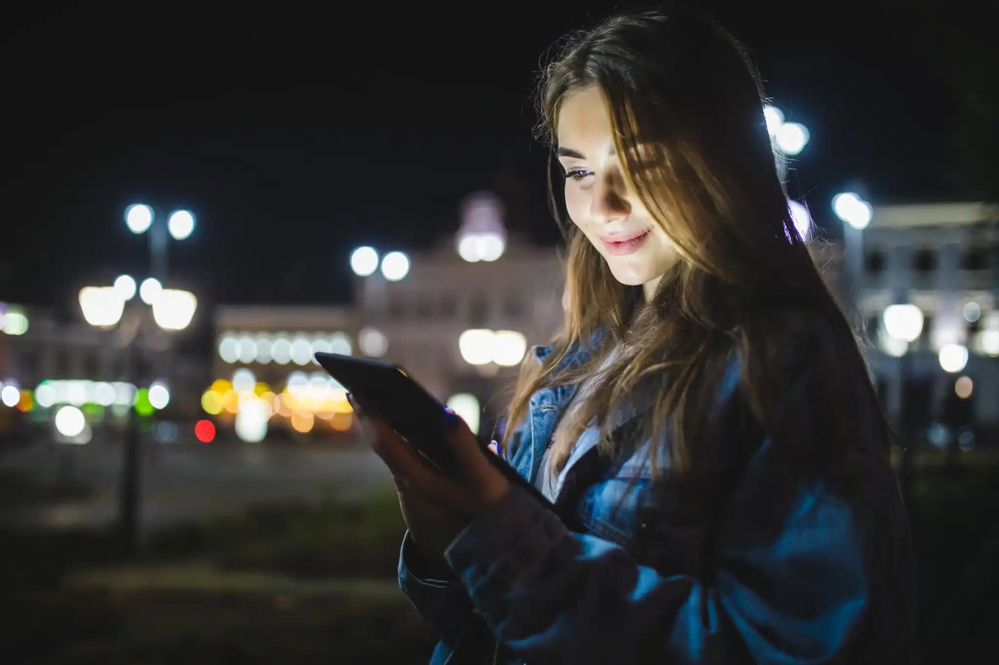 Mulher feliz usando celular sob as luzes da cidade à noite, em clima de boa noite.