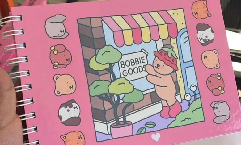 Imagem de um caderno com a capa mostrando uma ilustração fofa com uma loja chamada 'Bobbie Goods', um ursinho com chapéu de morango e vários personagens adoráveis ao redor
