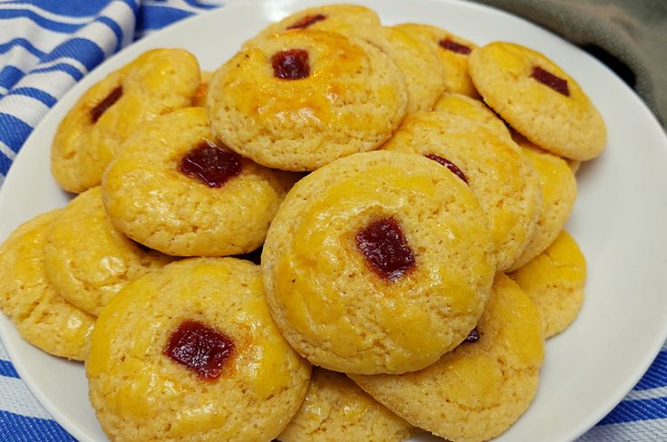 Broas de fubá douradas com pedaços de goiabada no centro, servidas em um prato branco, com toalha azul e branca ao fundo.