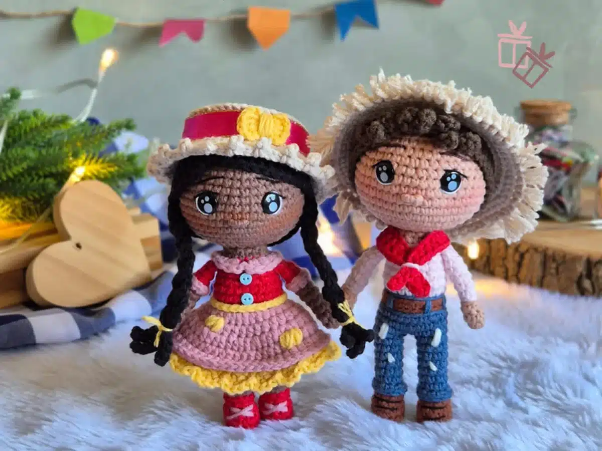 Casal de bonecos amigurumi em estilo junino com roupas típicas e decoração festiva ao fundo